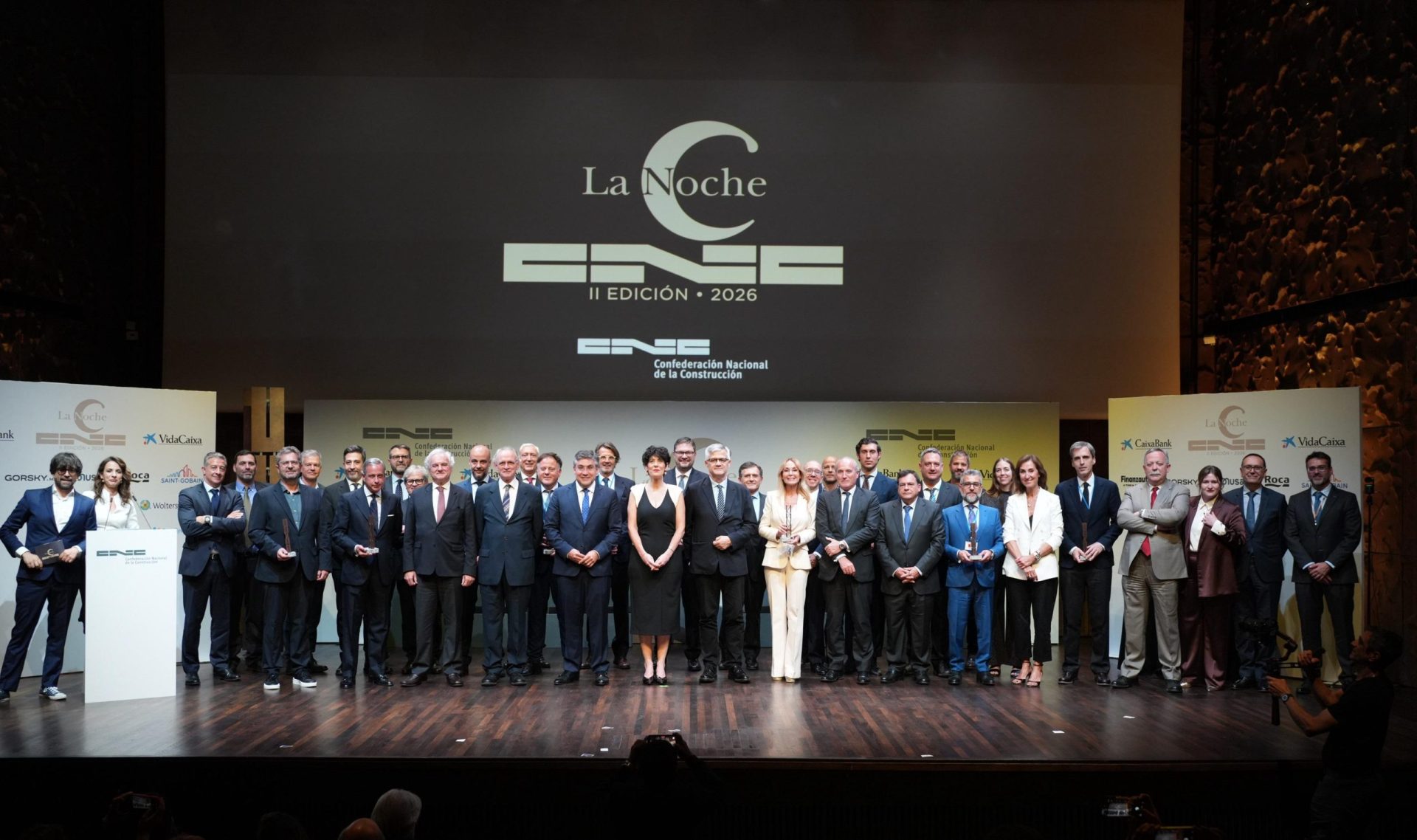 Group of professionals in formal attire posing on a stage at an awards ceremony, with a large screen displaying 'La Noche' and logos in the background.